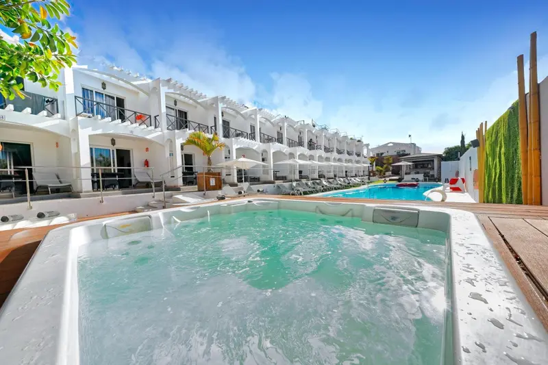 Jacuzzi en premier plan avec un bâtiment blanc et des transats près d'une piscine.
