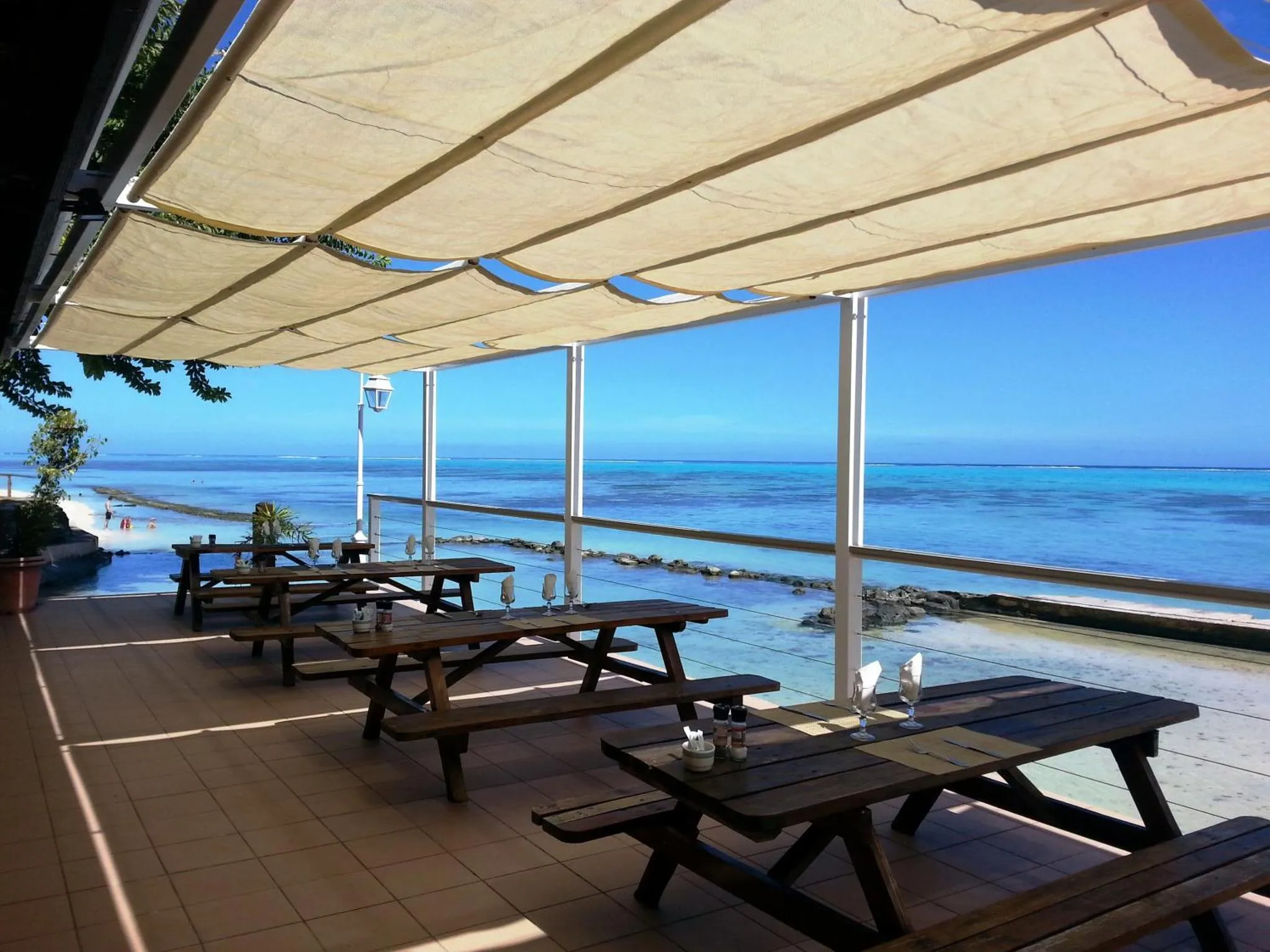Tables en bois disposées sur une terrasse ombragée face à la mer turquoise.
