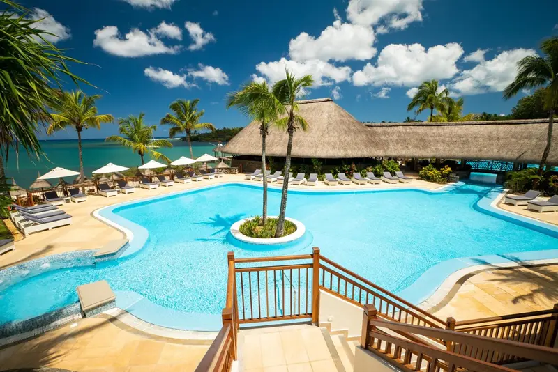 Une piscine turquoise bordée de transats et de palmiers sous un ciel bleu.