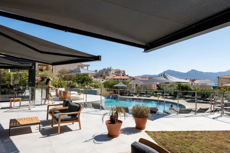 Terrasse ombragée avec mobilier d'extérieur donnant sur une piscine et un paysage côtier.
