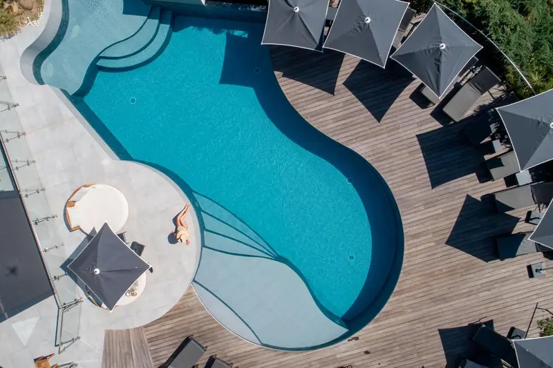 Vue aérienne d'une piscine rectangulaire entourée d'une terrasse en bois avec des parasols.