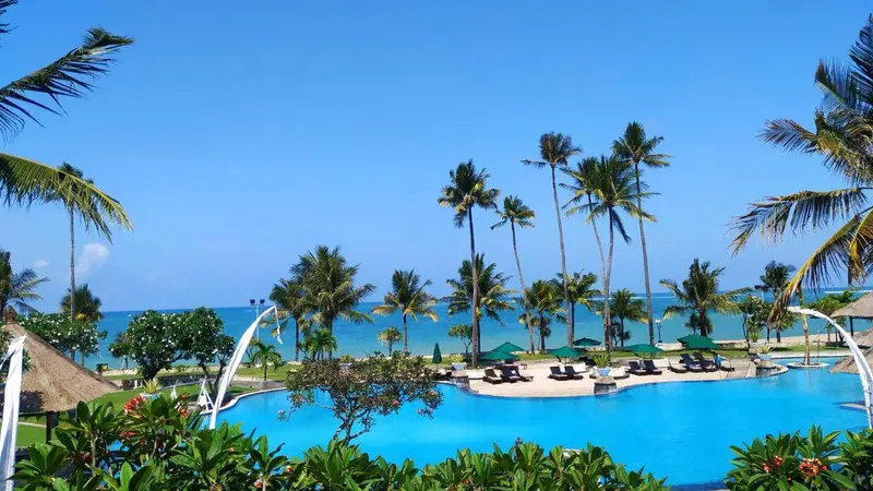 Une piscine bleue bordée de palmiers et de parasols, avec la mer en arrière-plan.