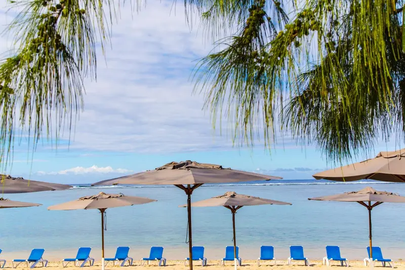 Transats bleus alignés sous des parasols sur une plage de sable fin, avec la mer turquoise à l'horizon.