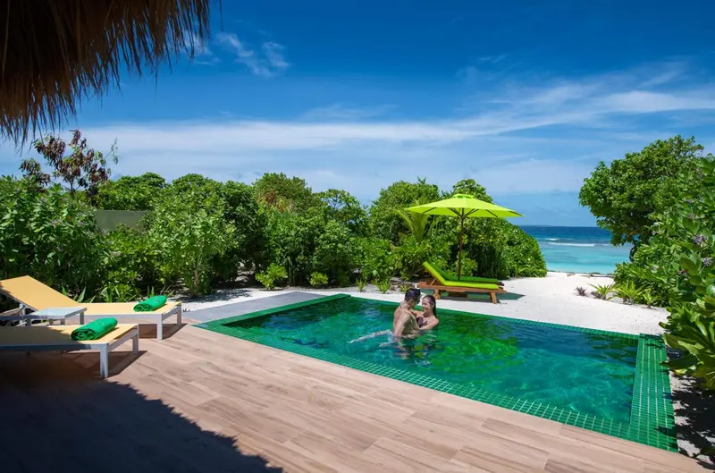 Couple se relaxant dans une piscine turquoise bordée de chaises longues sur une plage de sable blanc.