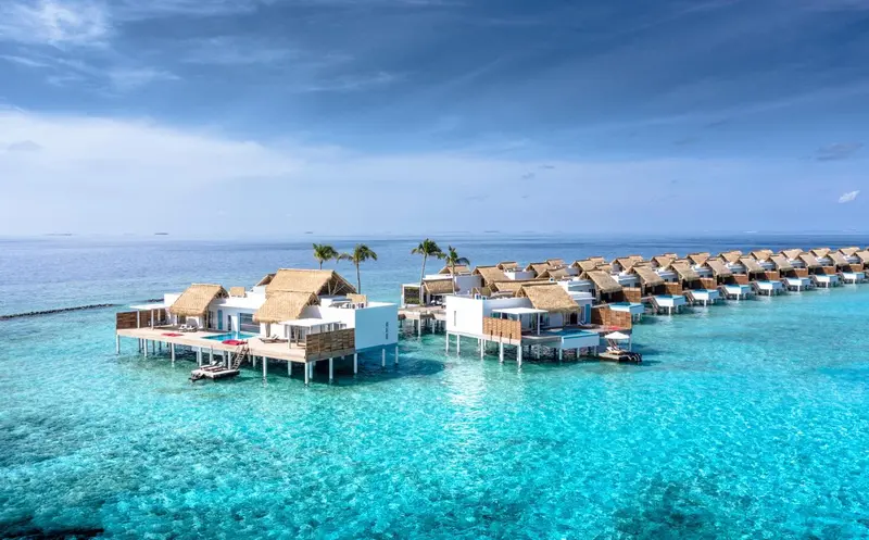 Vue aérienne d'un ensemble de bungalows sur pilotis au-dessus d'une eau turquoise.