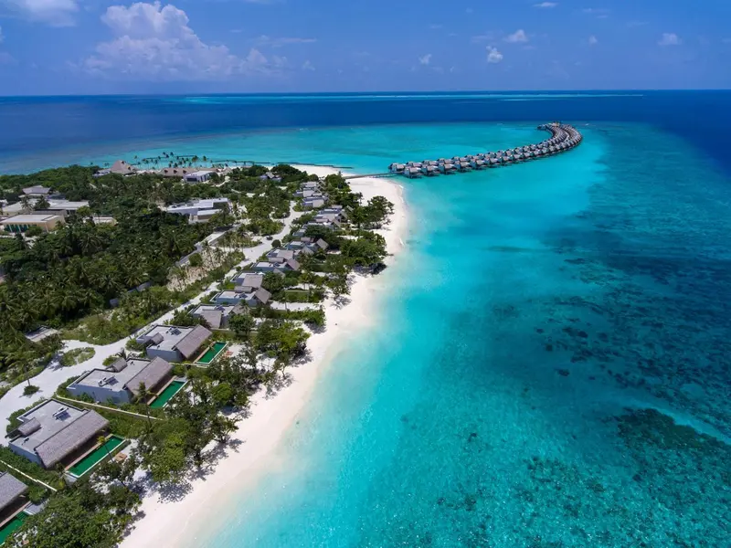 Vue aérienne d'une île tropicale avec des villas sur pilotis et une plage de sable blanc.