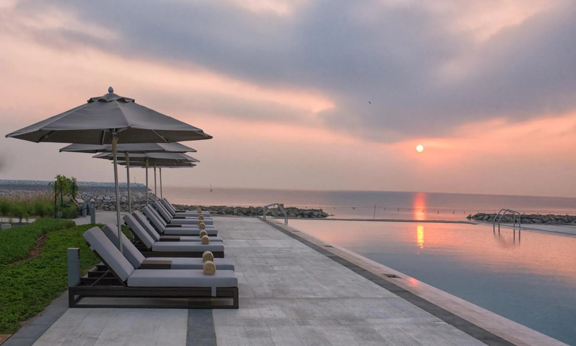 Transats alignés au bord d'une piscine à débordement face à la mer au coucher du soleil.