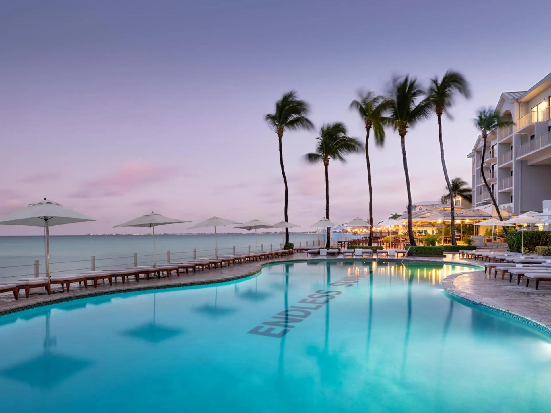 Une piscine bordée de chaises longues et de palmiers au coucher du soleil.