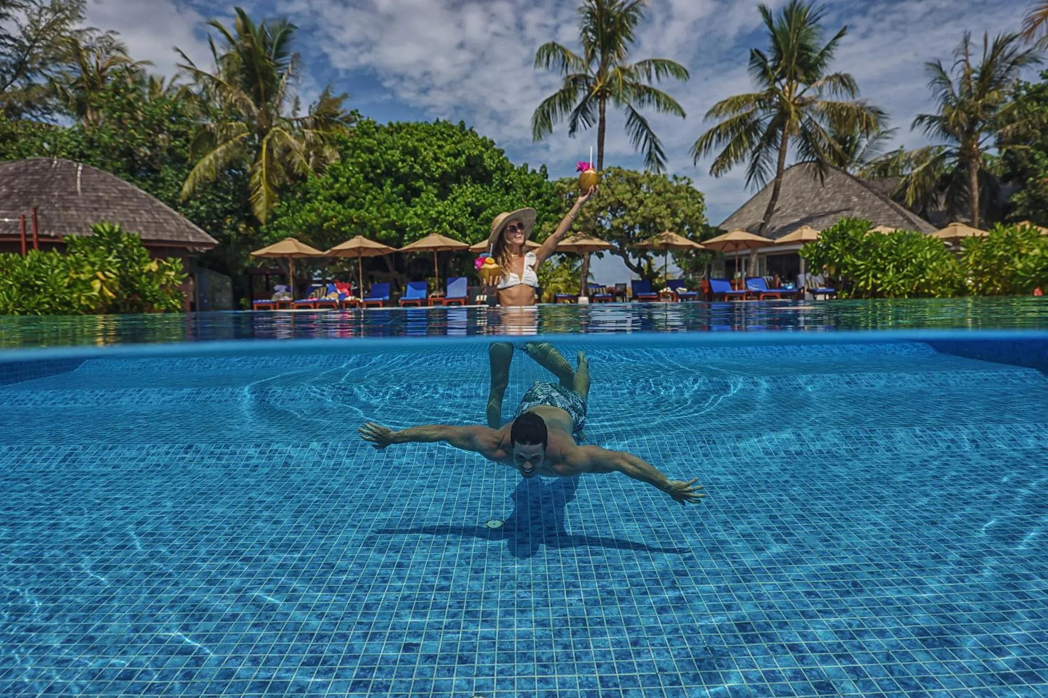 Une femme souriante lève un cocktail au-dessus d'une piscine où un homme nage.