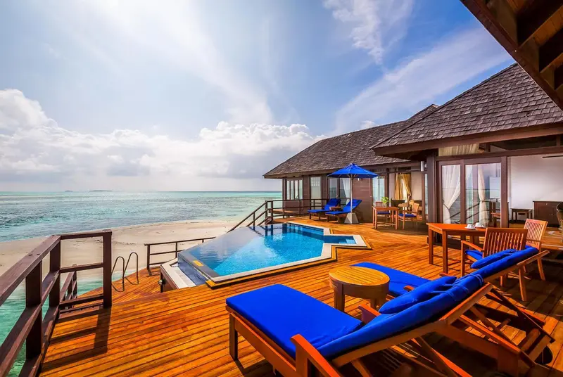 Terrasse en bois avec piscine et transats bleus donnant sur une plage de sable blanc et une mer turquoise.