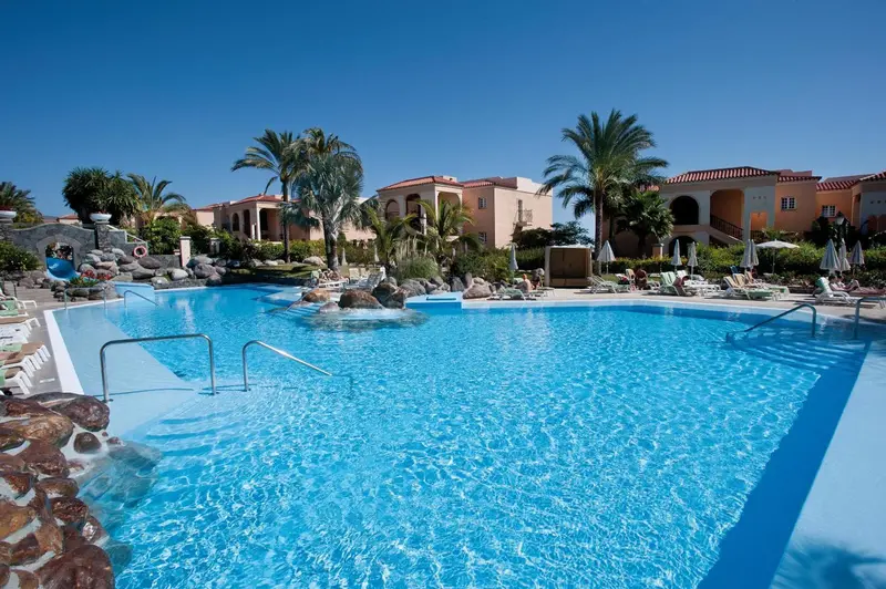 Une piscine turquoise bordée de transats et de rochers, avec des bâtiments aux toits rouges en arrière-plan.
