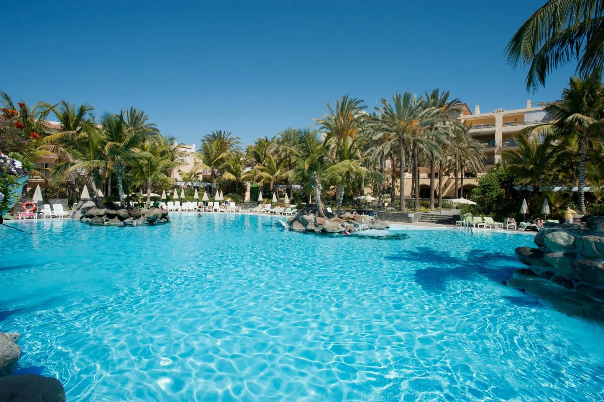 Une piscine turquoise bordée de rochers et de palmiers, avec des transats et des parasols.