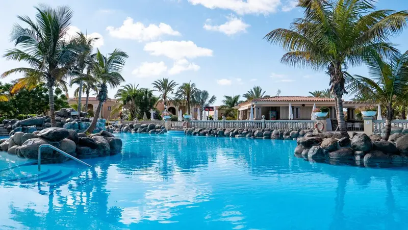 Une piscine turquoise bordée de rochers et de palmiers sous un ciel bleu.