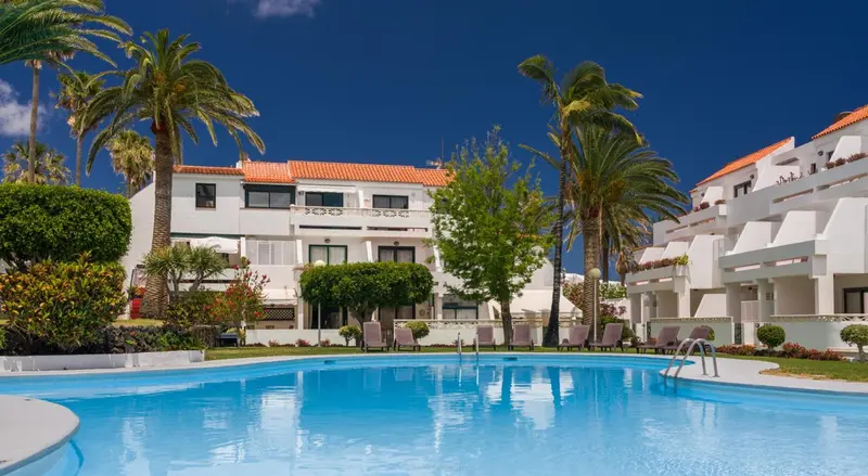 Vue d'une piscine turquoise devant un bâtiment blanc bordé de palmiers sous un ciel bleu.