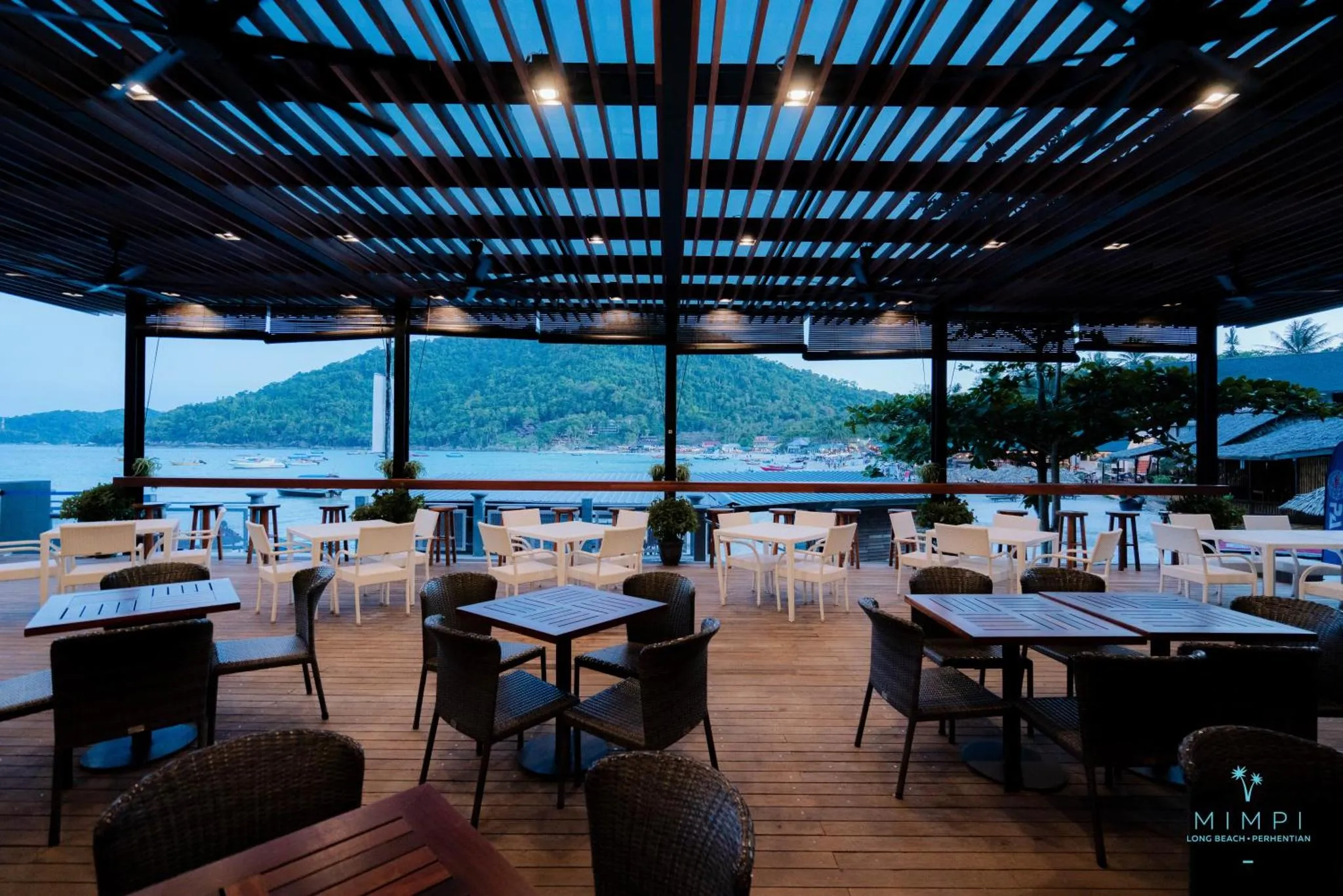 Terrasse ombragée avec tables et chaises donnant sur la mer et une île.