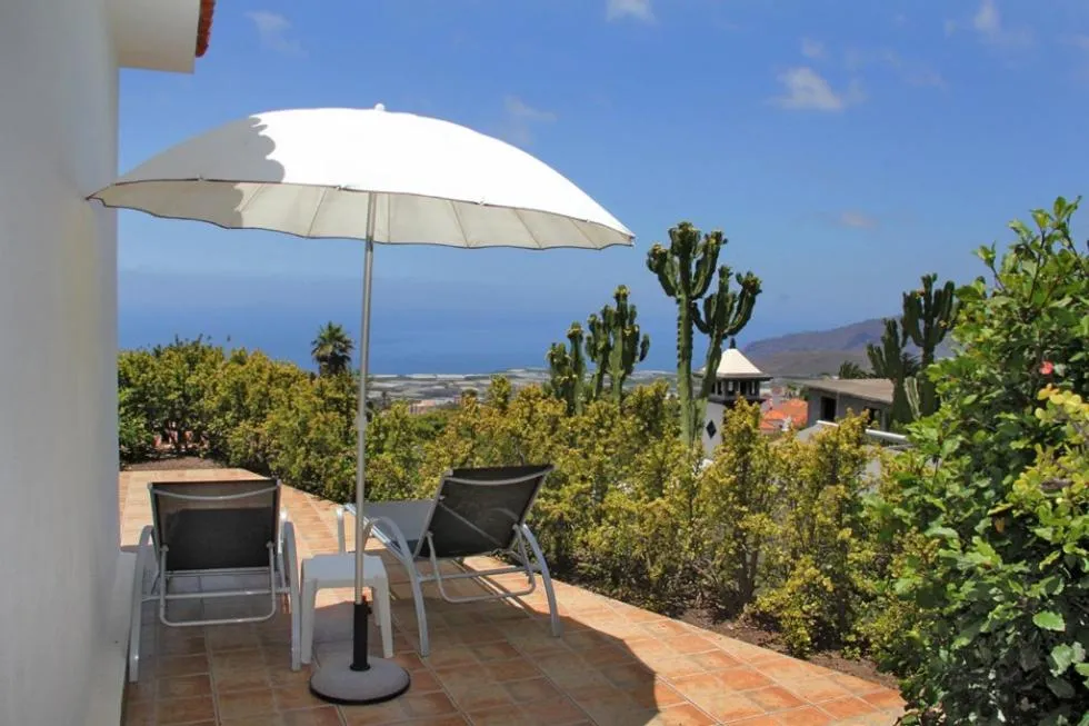 Deux transats blancs sous un parasol sur une terrasse carrelée, avec une vue sur la mer et la végétation.