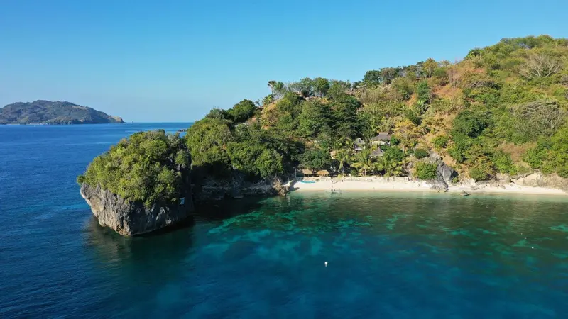 Vue aérienne d'une plage de sable blanc bordée de végétation luxuriante et de falaises rocheuses.