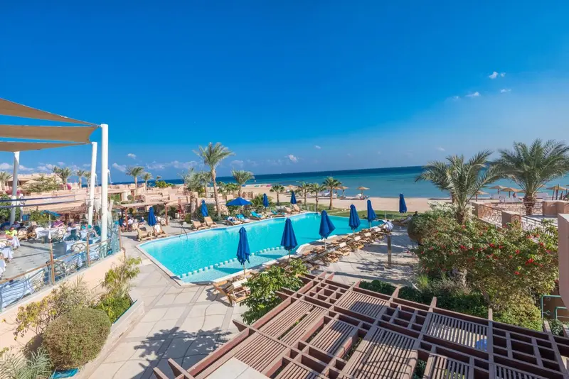 Vue aérienne d'une piscine bordée de transats et de parasols, avec la mer en arrière-plan.