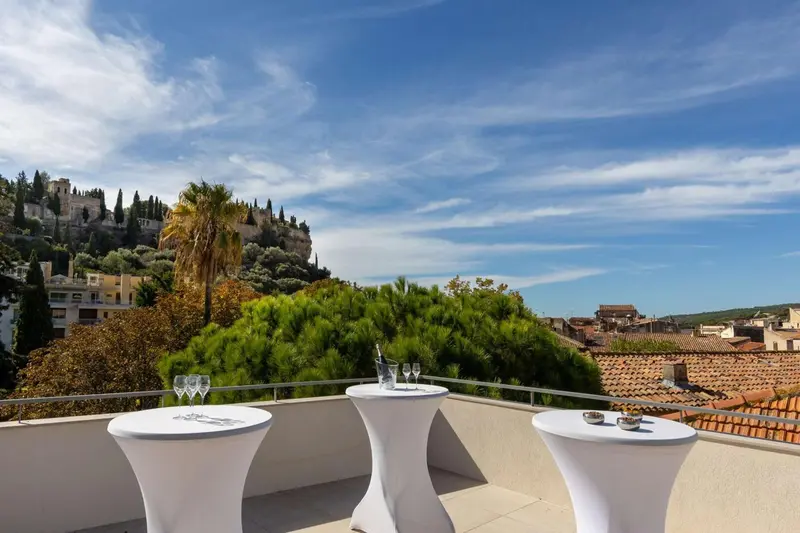 Tables rondes blanches sur une terrasse ensoleillée avec vue sur la ville et la végétation.