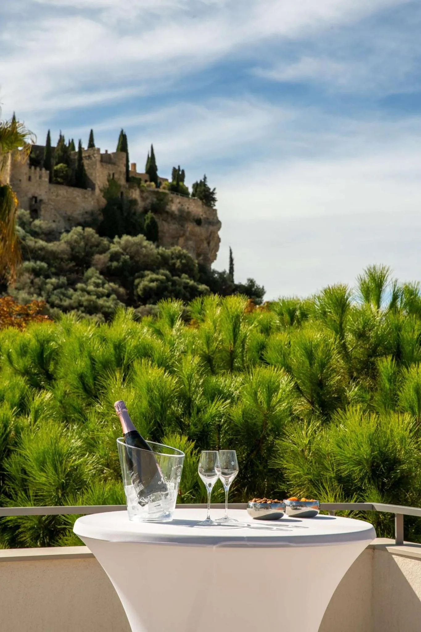 Une table avec une bouteille de champagne et des verres est placée sur une terrasse ensoleillée avec vue sur un paysage verdoyant et un château.
