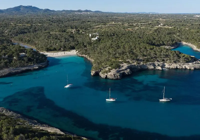 Vue aérienne d'une crique turquoise bordée de végétation et de quelques voiliers.