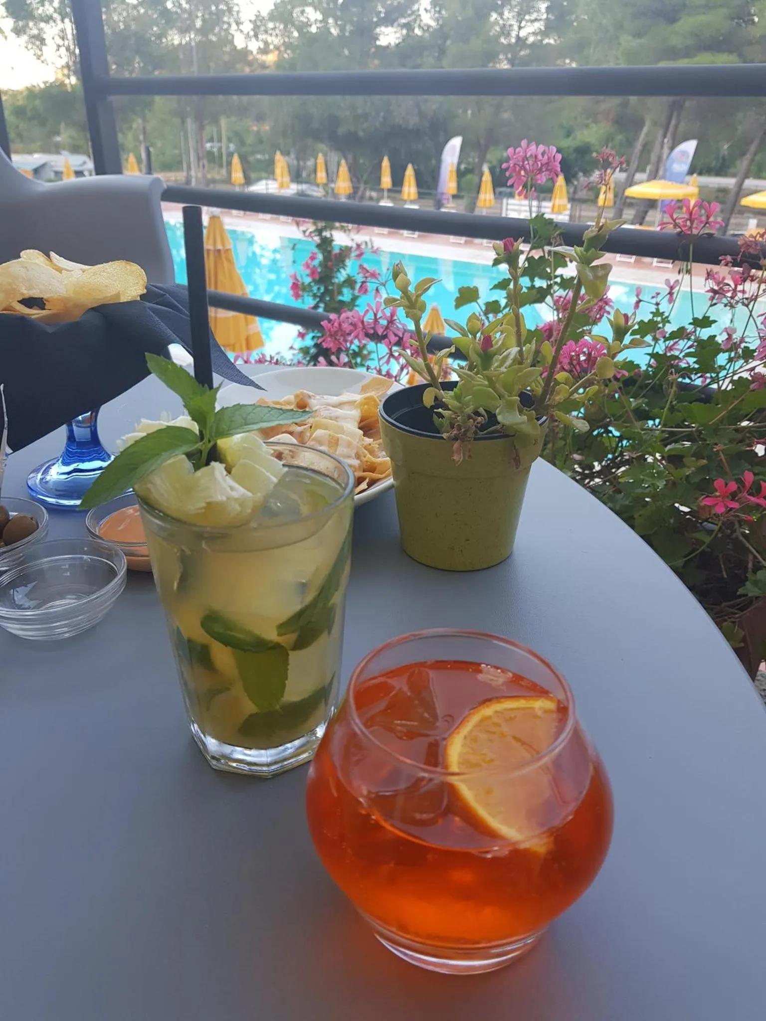 Une table d'extérieur présente des boissons et des en-cas devant une piscine ensoleillée.