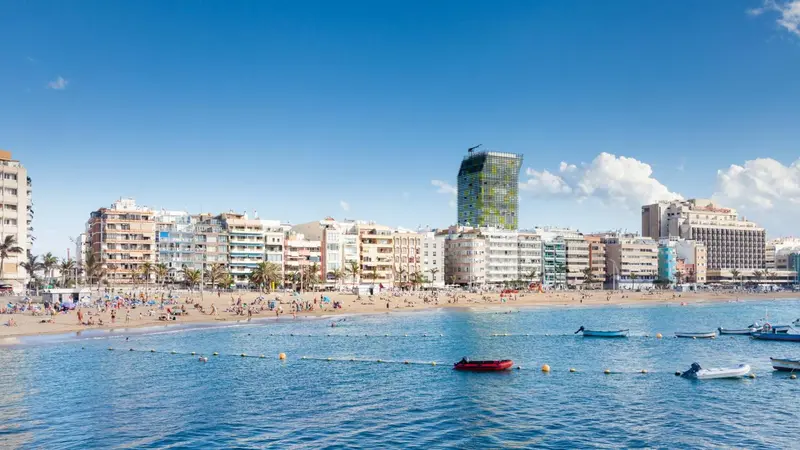 Vue d'une plage ensoleillée avec des bâtiments en arrière-plan et des bateaux sur l'eau.