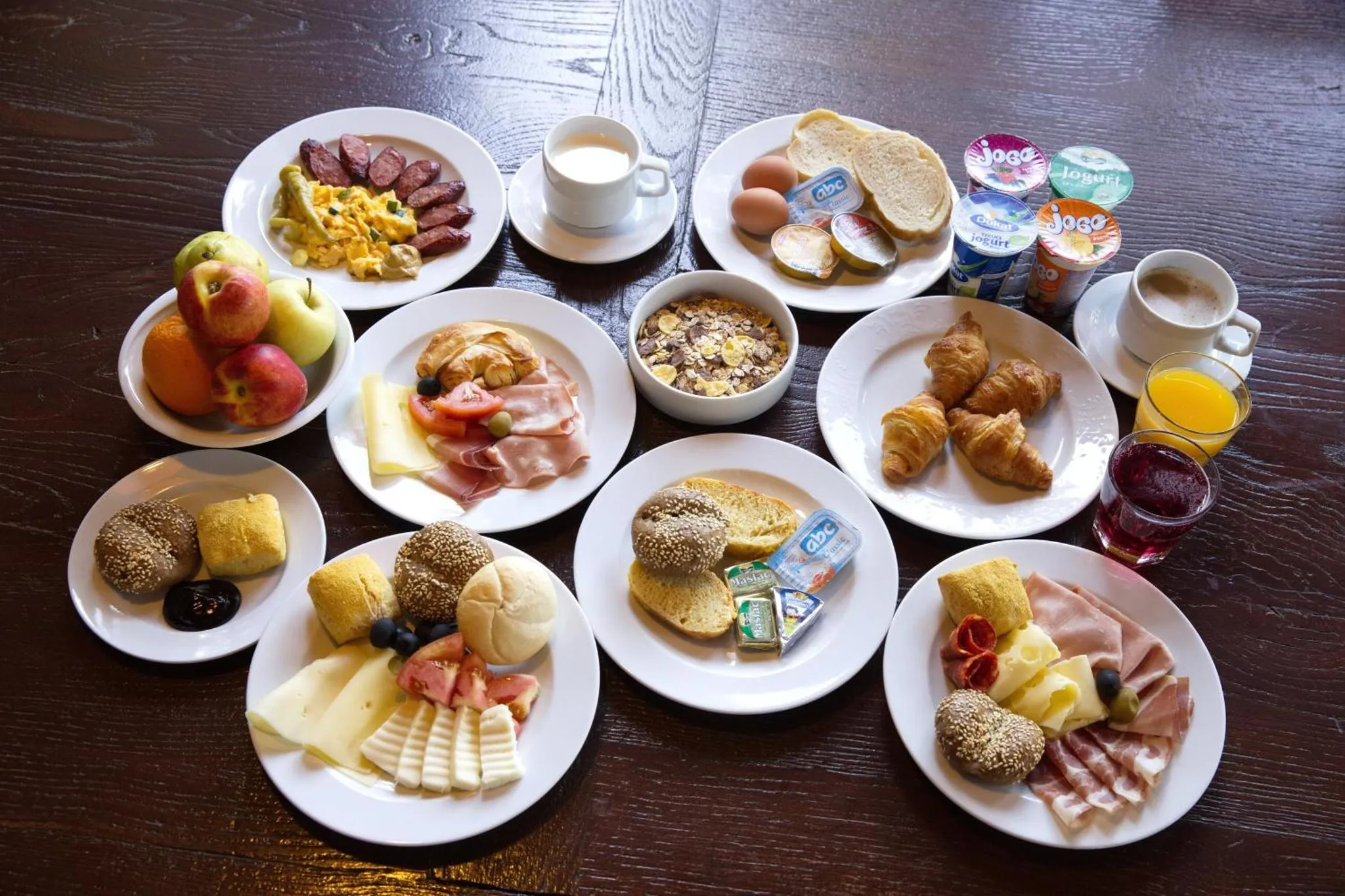 Une table est dressée avec un copieux petit-déjeuner composé de viennoiseries, de charcuteries, de fruits et de boissons.