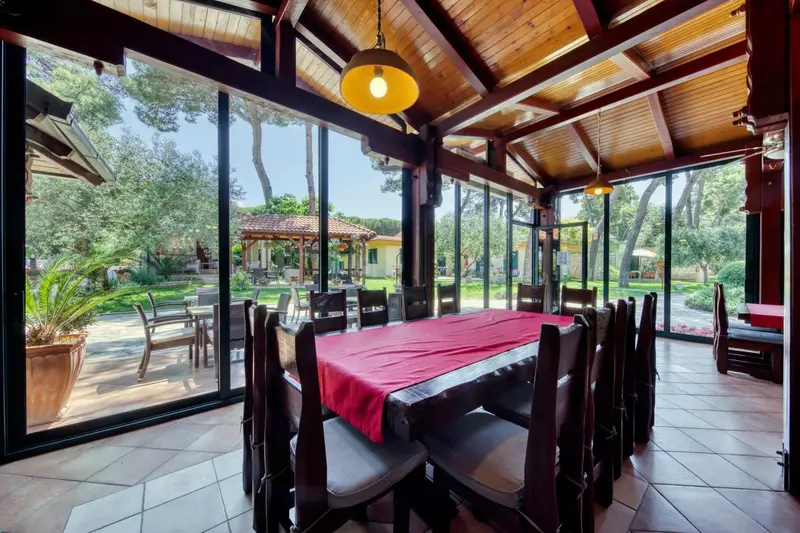 Une grande table à manger en bois est dressée dans une salle lumineuse avec de larges baies vitrées.