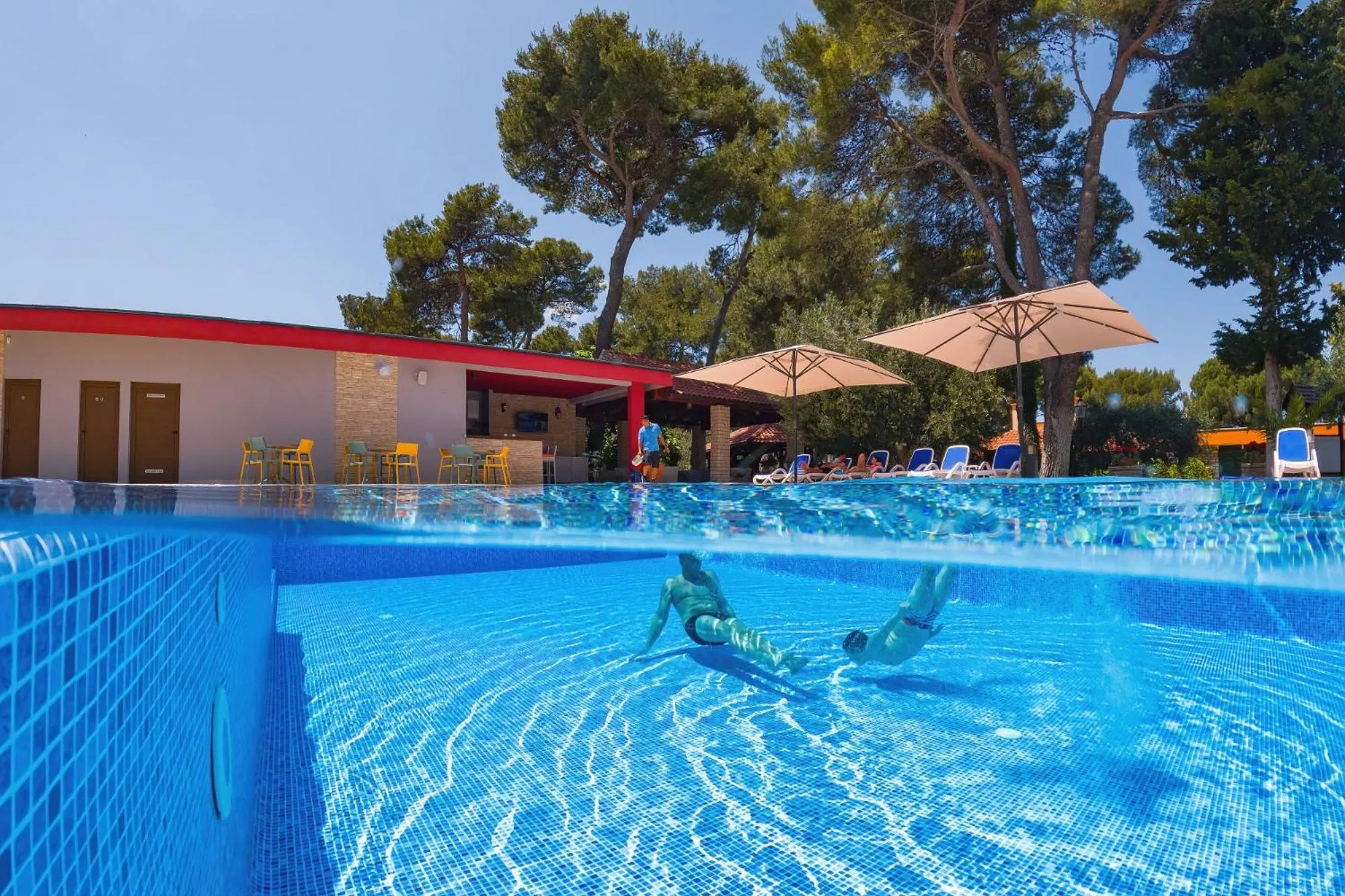 Deux personnes nagent dans une piscine ensoleillée avec des transats et des parasols à proximité.