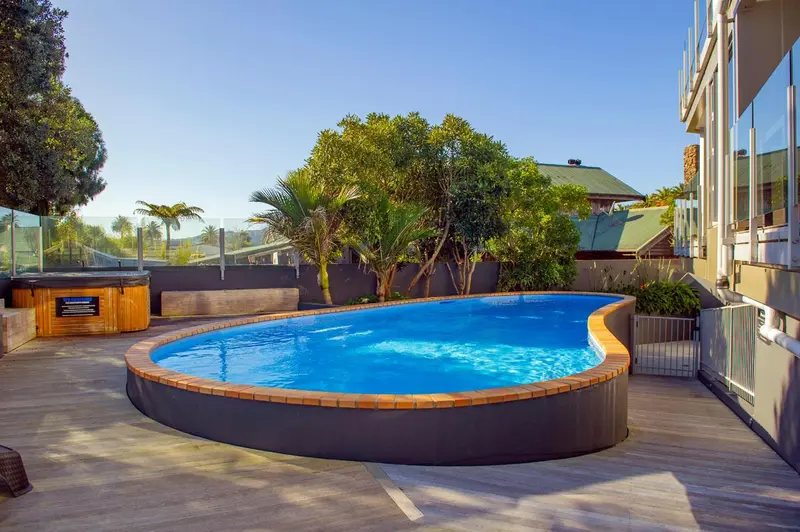 Une piscine rectangulaire bordée de briques est entourée d'une terrasse en bois et de végétation luxuriante.