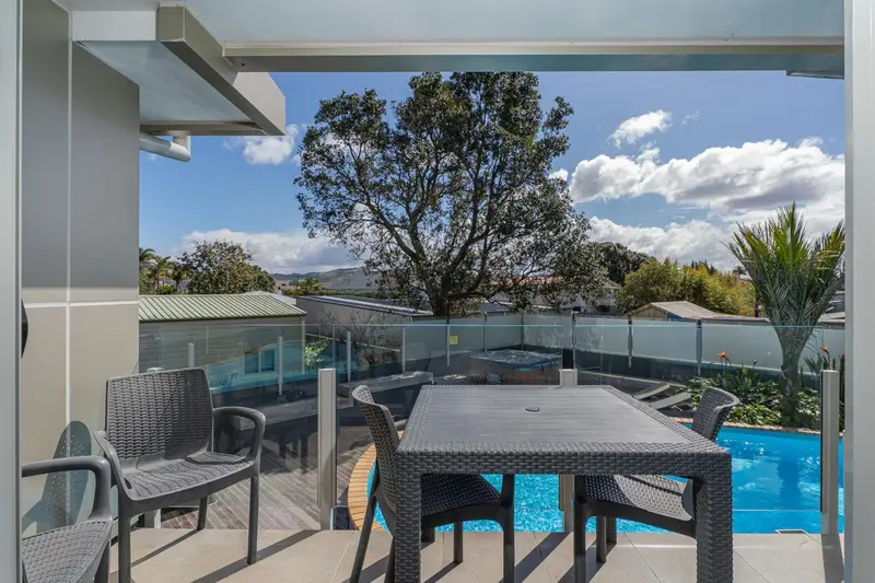 Une table et des chaises en osier sont disposées sur une terrasse carrelée avec vue sur une piscine et un paysage verdoyant.
