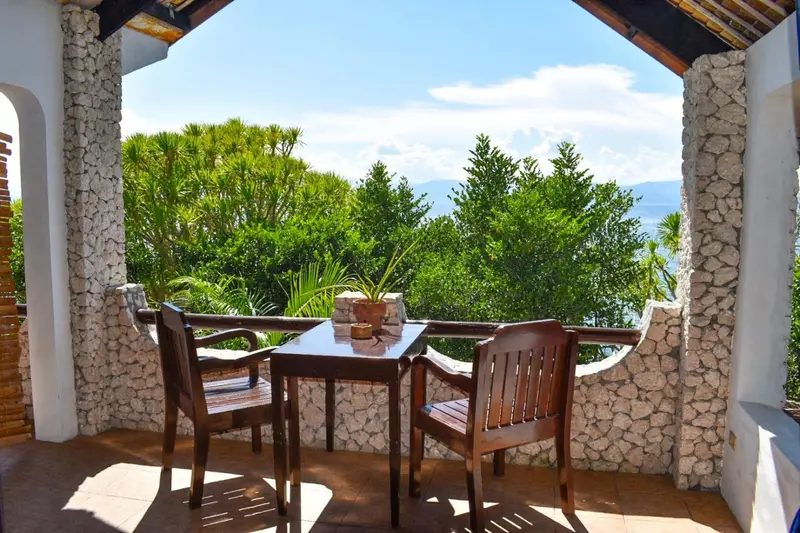 Une table et deux chaises en bois sont disposées sur une terrasse ombragée avec vue sur la végétation et la mer.