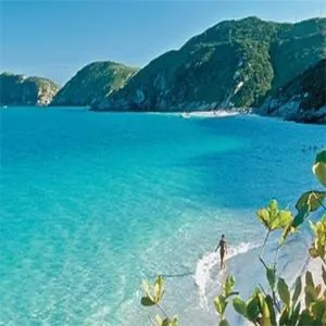 Une personne se tient dans l'eau turquoise d'une plage de sable clair, avec des collines verdoyantes en arrière-plan.