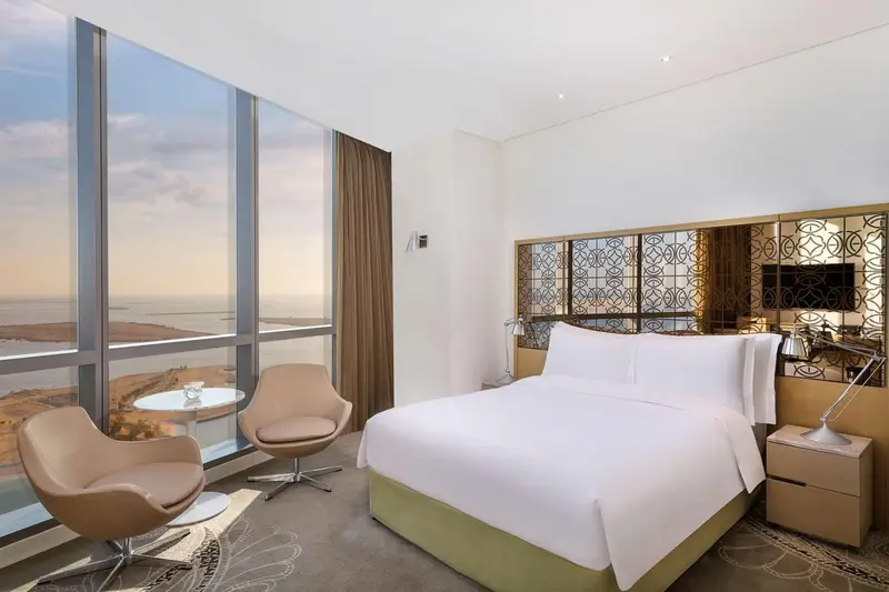 Chambre lumineuse avec un grand lit et un coin salon près d'une fenêtre panoramique.