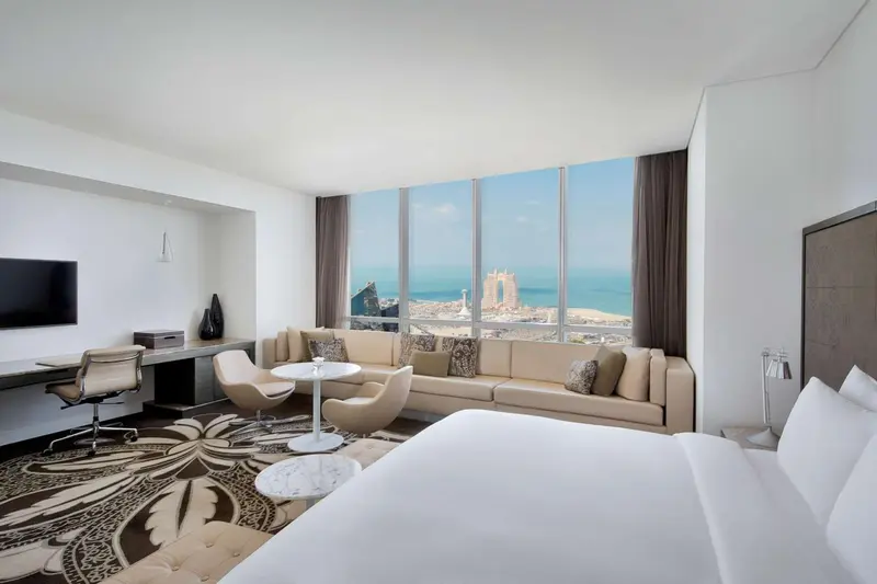 Une chambre lumineuse avec un grand lit et un coin salon donnant sur une vue urbaine et maritime.