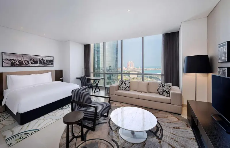Une chambre lumineuse avec un grand lit et un coin salon donnant sur une vue urbaine.