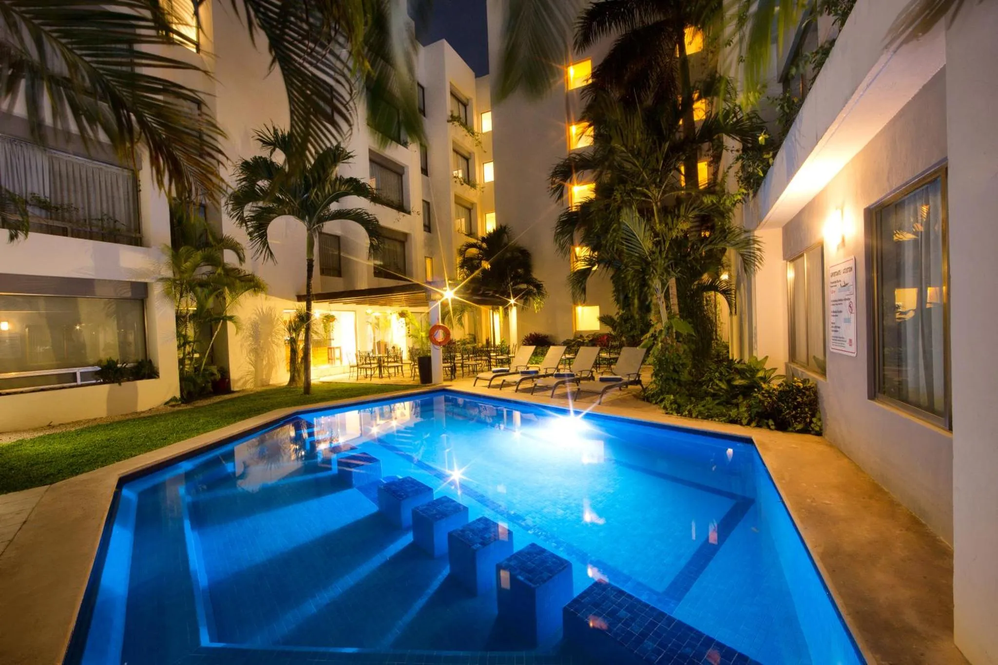Piscine illuminée dans une cour intérieure d'hôtel avec végétation tropicale.