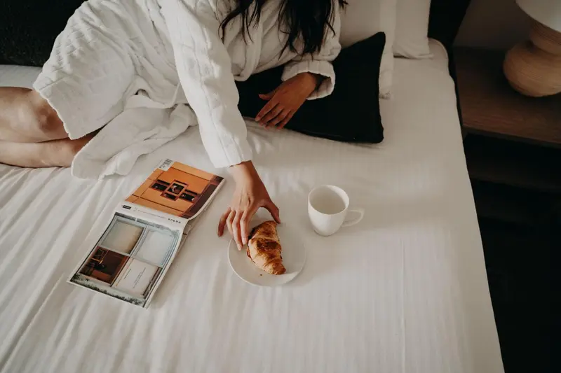 Une personne est assise sur un lit blanc avec un croissant et une tasse de café.