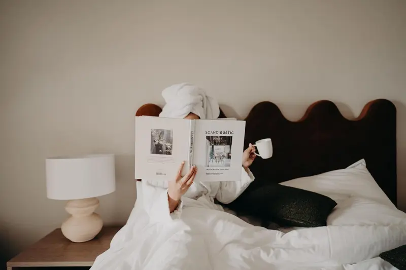 Une personne lit un livre au lit dans une chambre lumineuse.