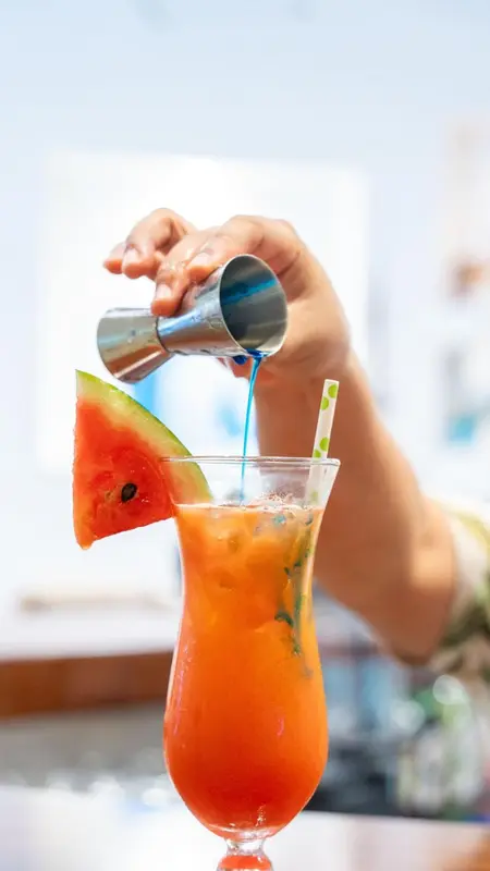 Une main verse un liquide bleu dans un cocktail à la pastèque.