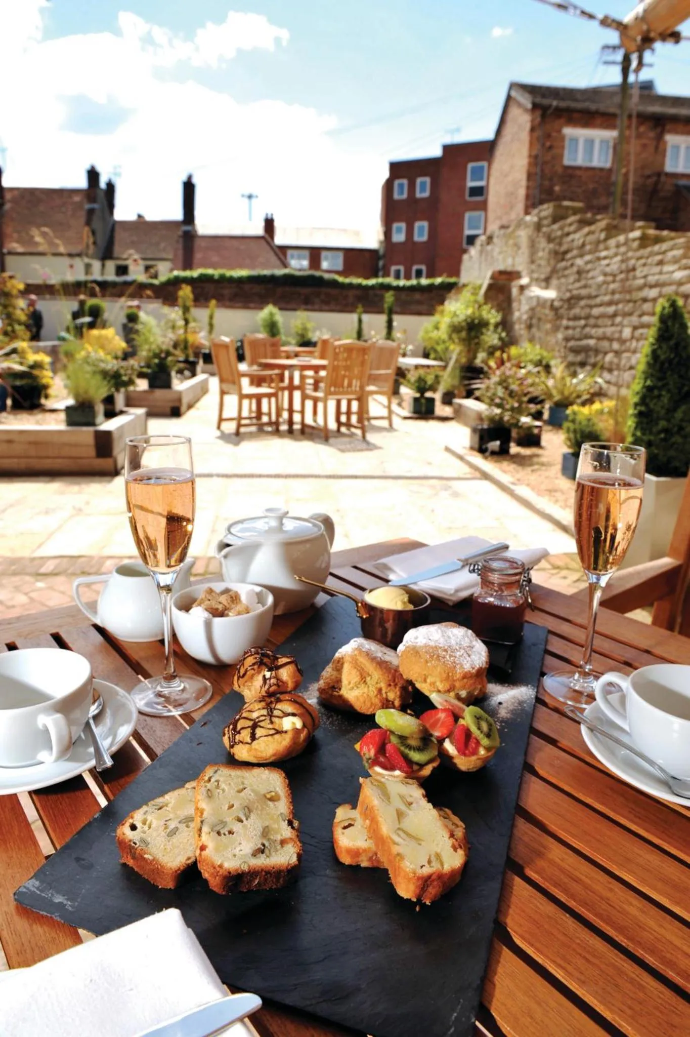 Une table est dressée avec des pâtisseries, des boissons et de la confiture sur une terrasse pavée.
