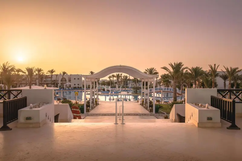 Une allée mène vers une piscine au sein d'un complexe hôtelier tropical au coucher du soleil.