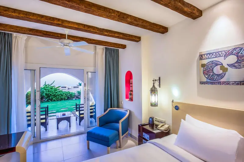 Une chambre lumineuse avec un lit blanc et un fauteuil bleu, donnant sur une terrasse verdoyante.