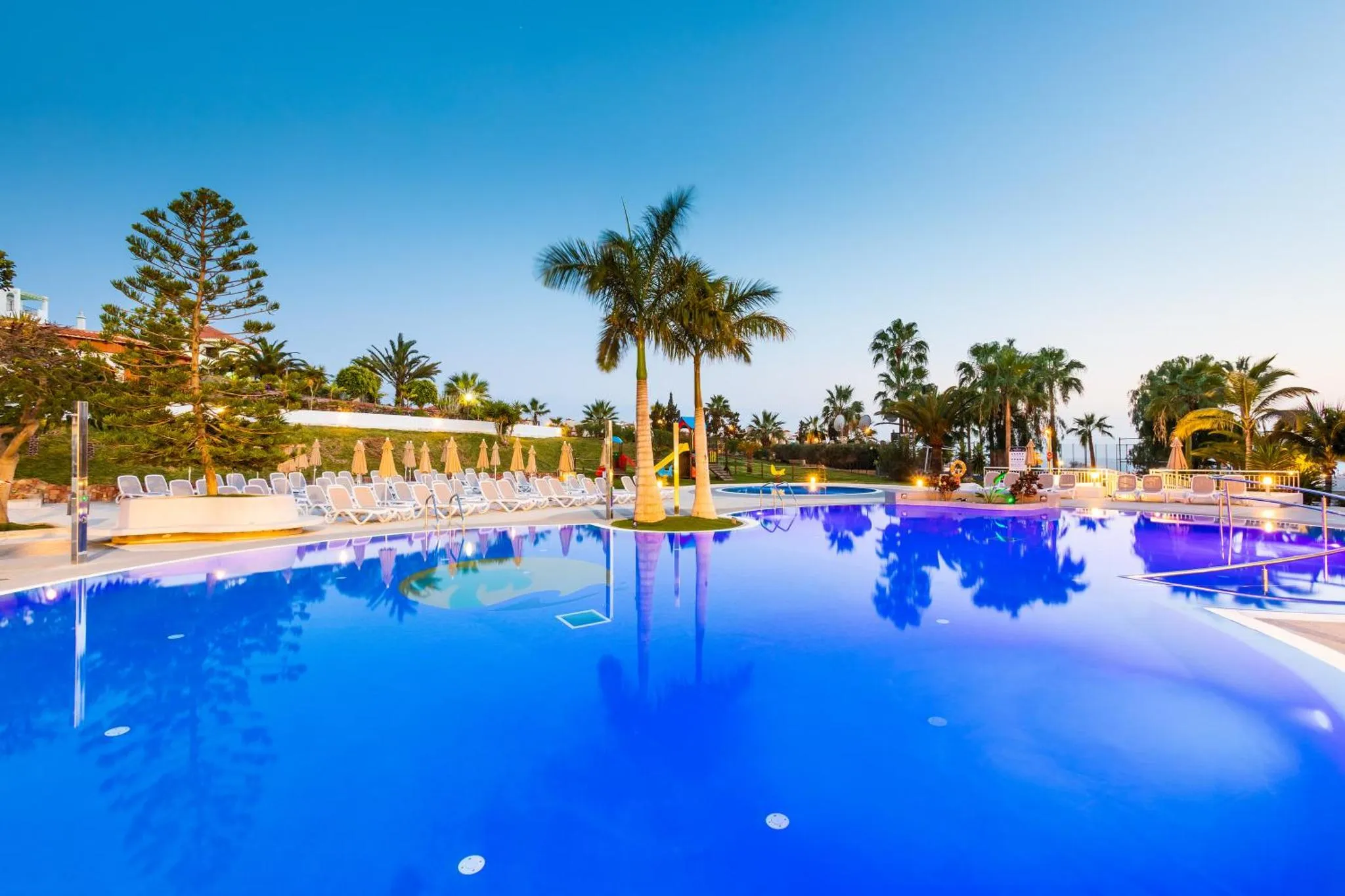 Piscine illuminée avec transats et palmiers sous un ciel bleu.