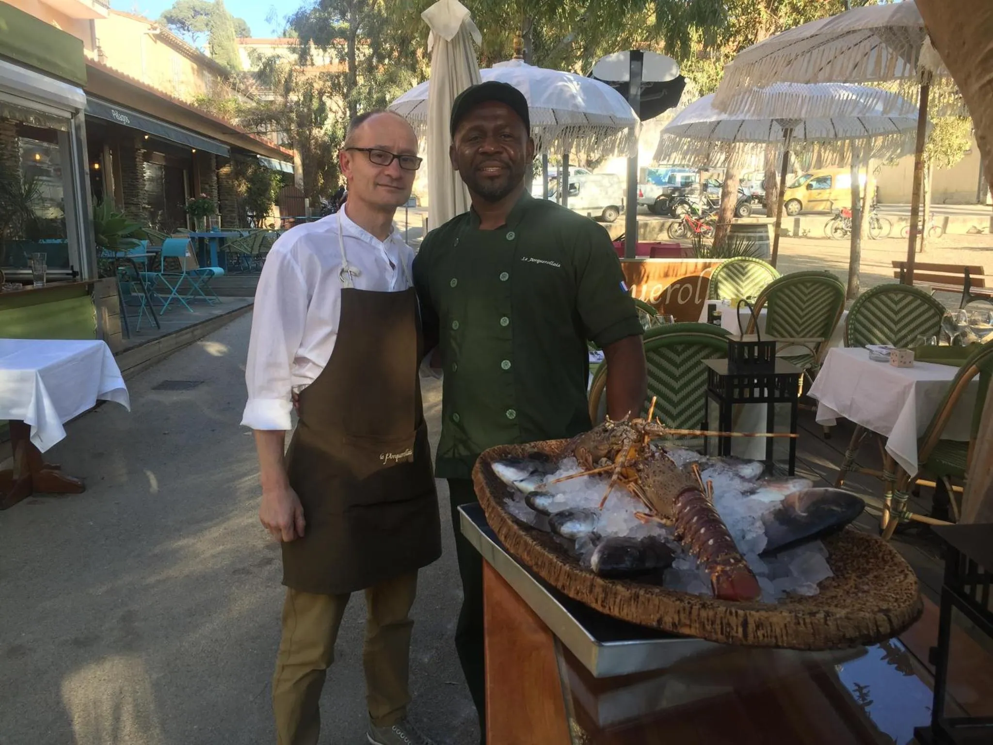 Deux hommes souriants, en tenue de cuisine, présentent un plateau de fruits de mer frais sur une terrasse.