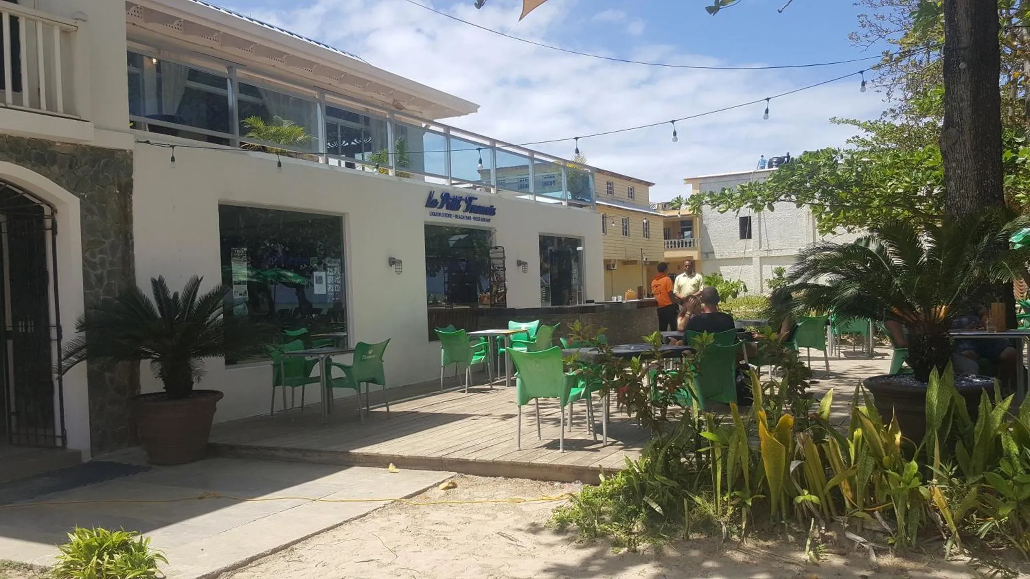 Terrasse ombragée avec tables et chaises vertes devant un bâtiment blanc.