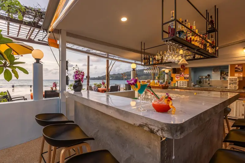 Un comptoir de bar en béton clair présente des boissons colorées, face à une vue sur la mer.