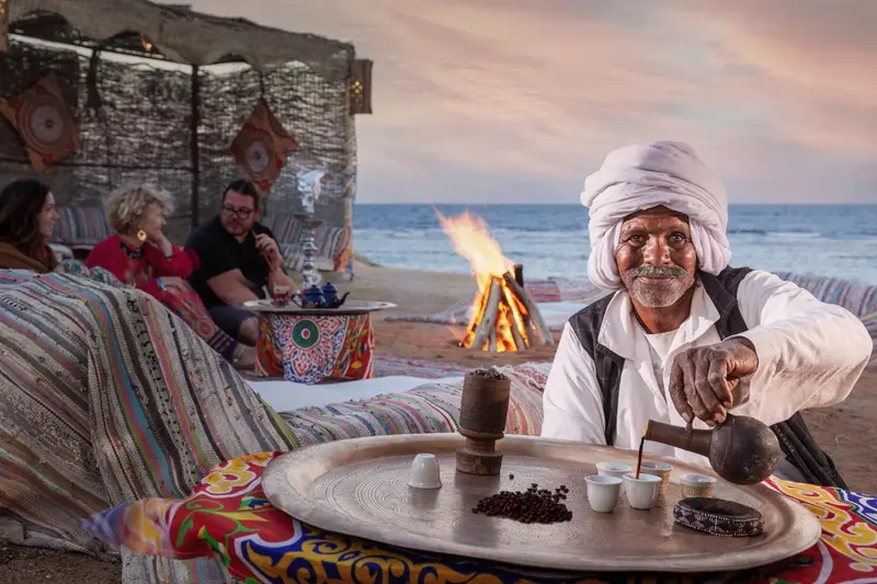 Un homme prépare du café sur le sable près d'un feu de bois.