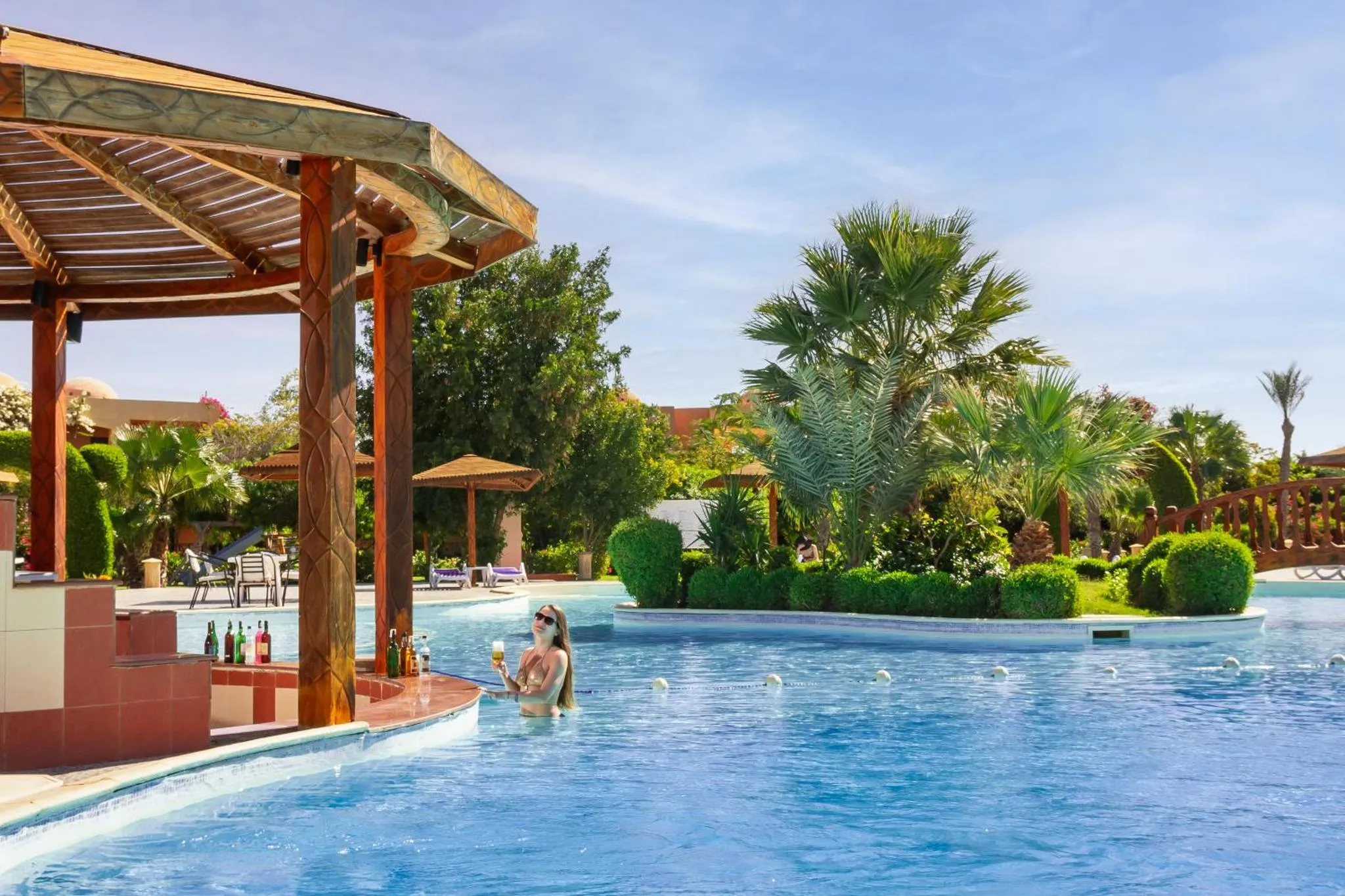 Une personne se tient dans une piscine avec un verre à la main, près d'un bar extérieur.